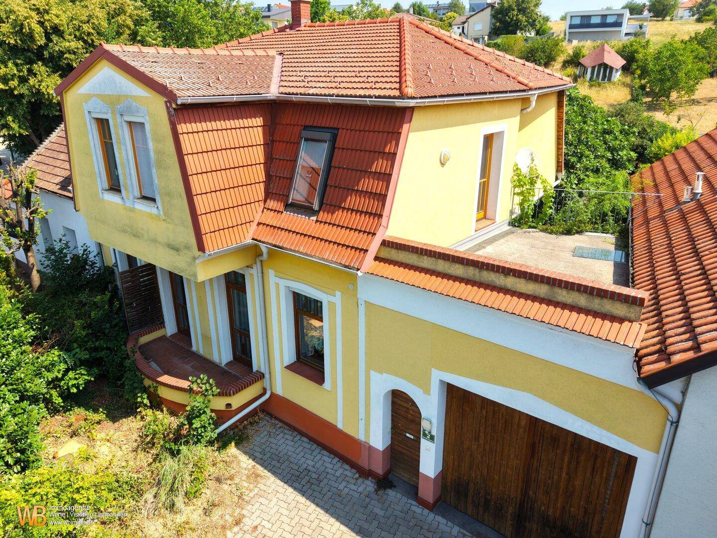 Gelbes Haus mit Mansarddach, rotem Ziegeldach und integrierter Garage, umgeben von üppigem Grün.