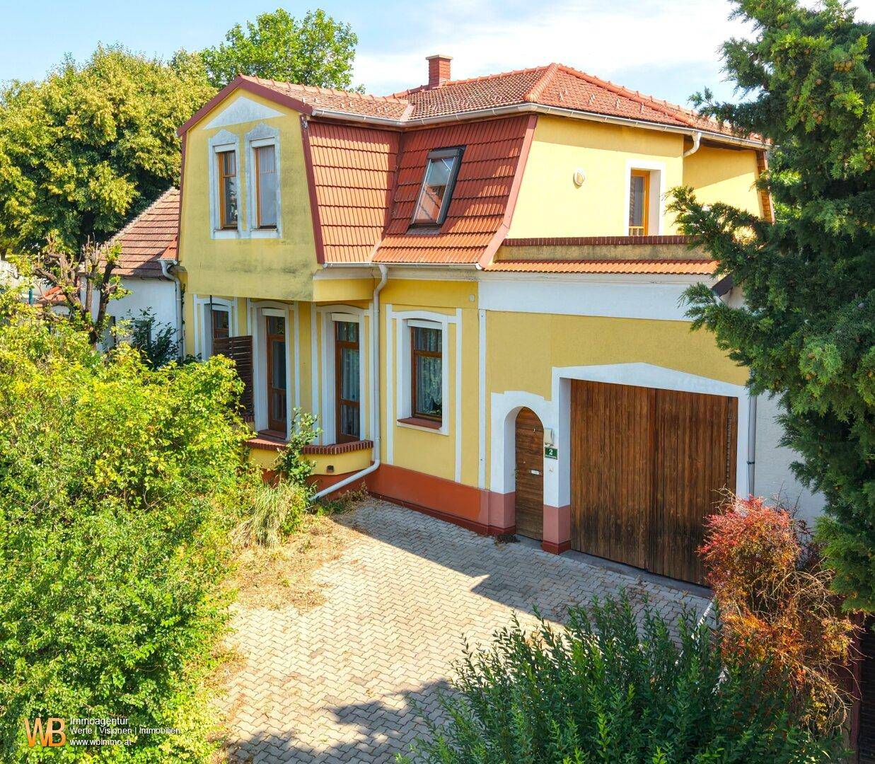 Vorderansicht des gelben Hauses mit Mansarddach, Holztor der Garage und gepflasterter Einfahrt.