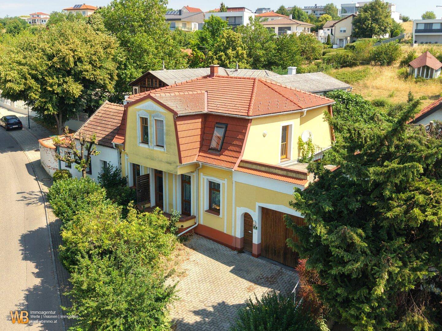 Weitläufige Ansicht des gelben Hauses mit rotem Dach, umgeben von Bäumen und einer ruhigen Straße.