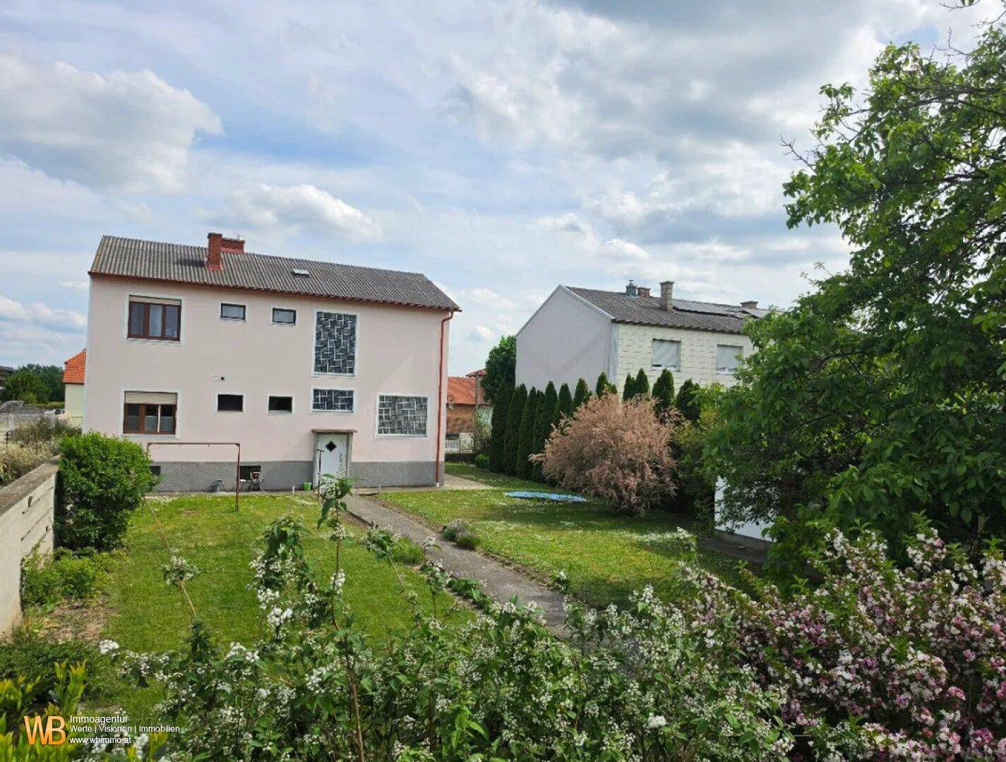 Blick auf den hinteren Gartenbereich mit Rasenfläche, Bäumen und einem weiteren Gebäude im Hintergrund.