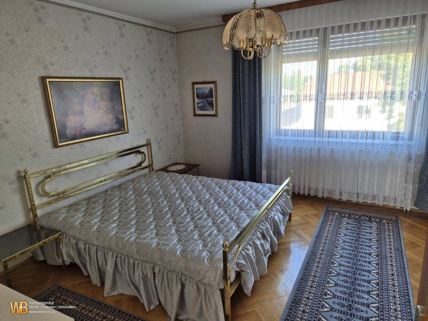 Schlafzimmer mit Messingbett, Parkettboden und großen Fenstern, die viel Licht hereinlassen.
