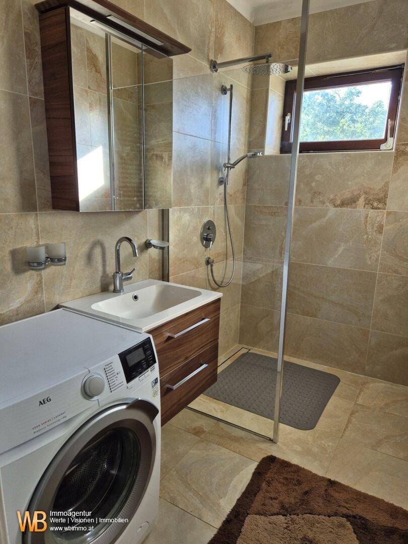 Modernisiertes Badezimmer mit beigefarbenen Fliesen, ebenerdiger Dusche und Waschmaschine.