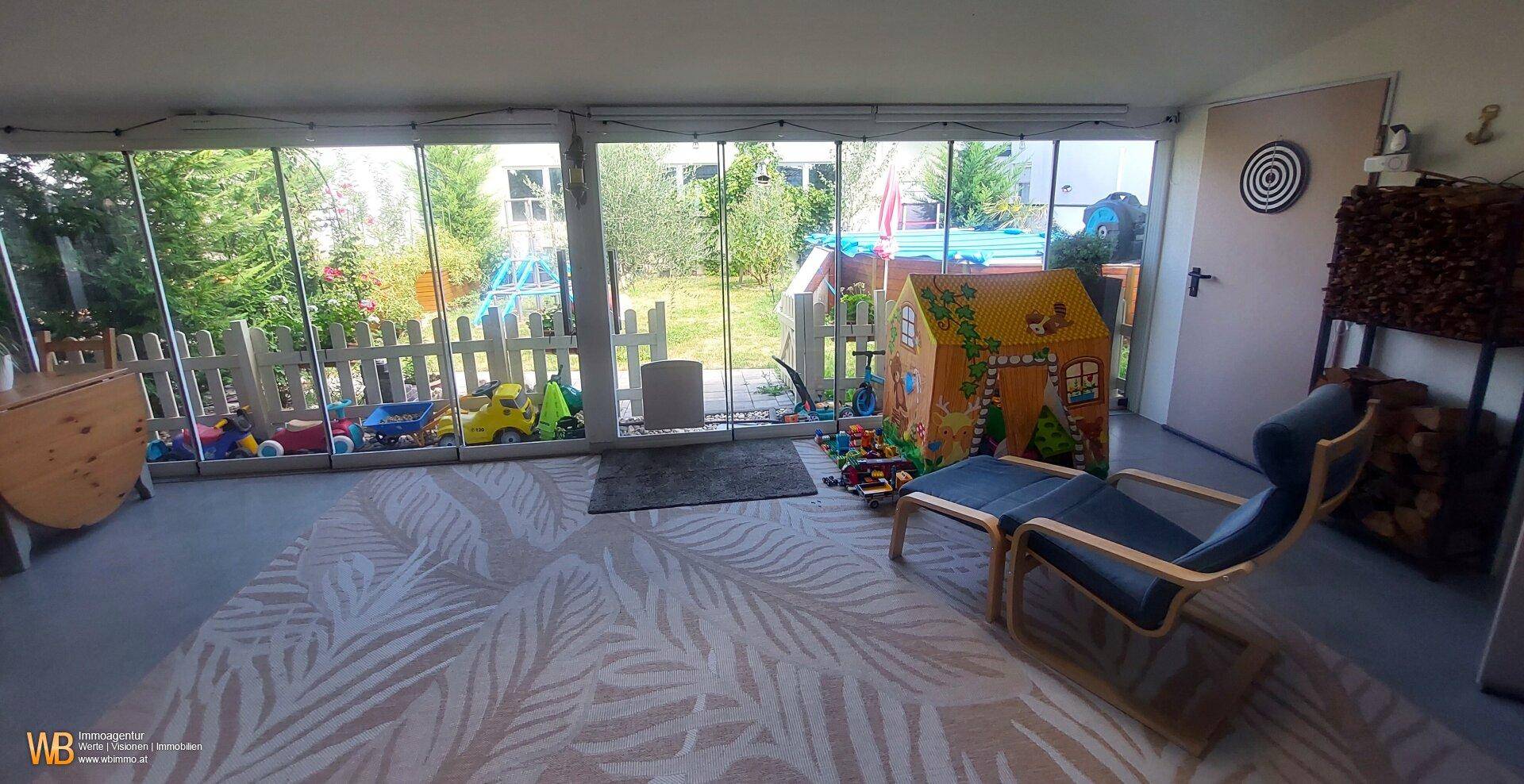 Heller Wintergarten mit großen Fenstern, Teppichboden und Blick auf den Spielbereich im Garten.