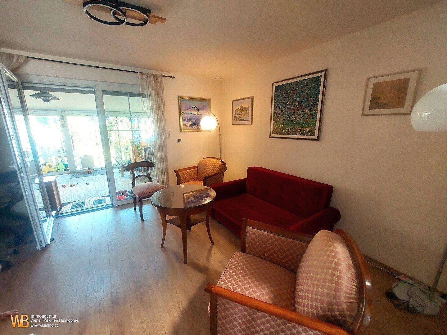 Gemütliches Wohnzimmer mit rotem Sofa, klassischen Sesseln und Zugang zu einem hellen Außenbereich.