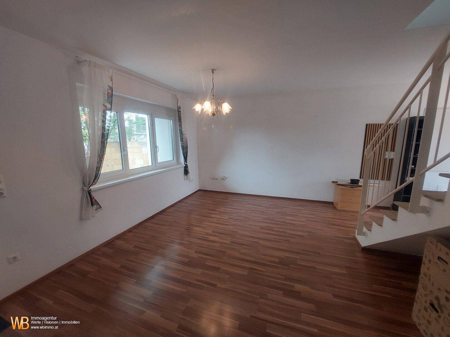 Heller Wohnbereich mit Holzfußboden, großem Fenster und Zugang zur Treppe.