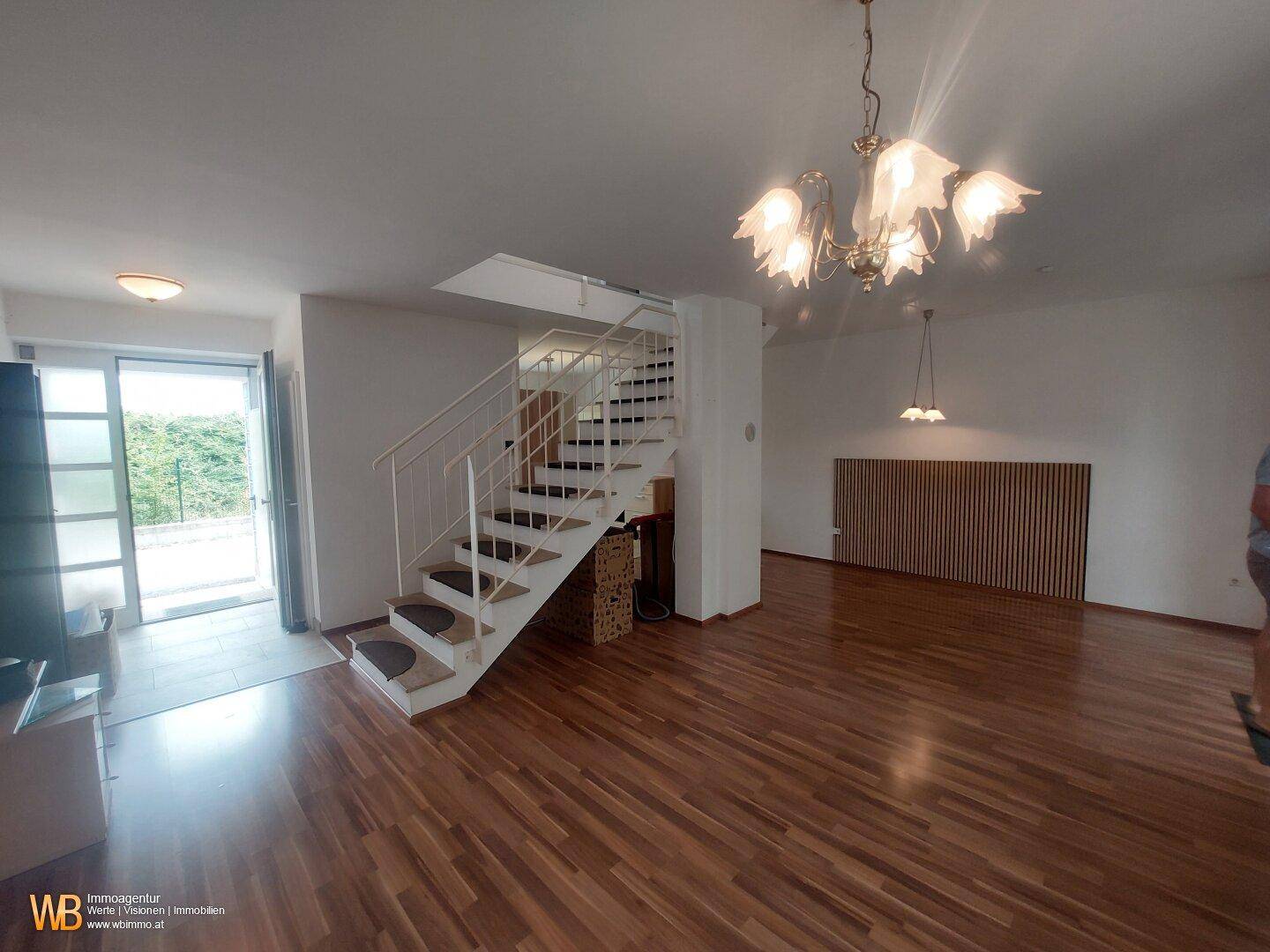 Geräumiges Wohnzimmer mit Holzparkettboden, offener Treppe und Zugang zu einem Außenbereich.