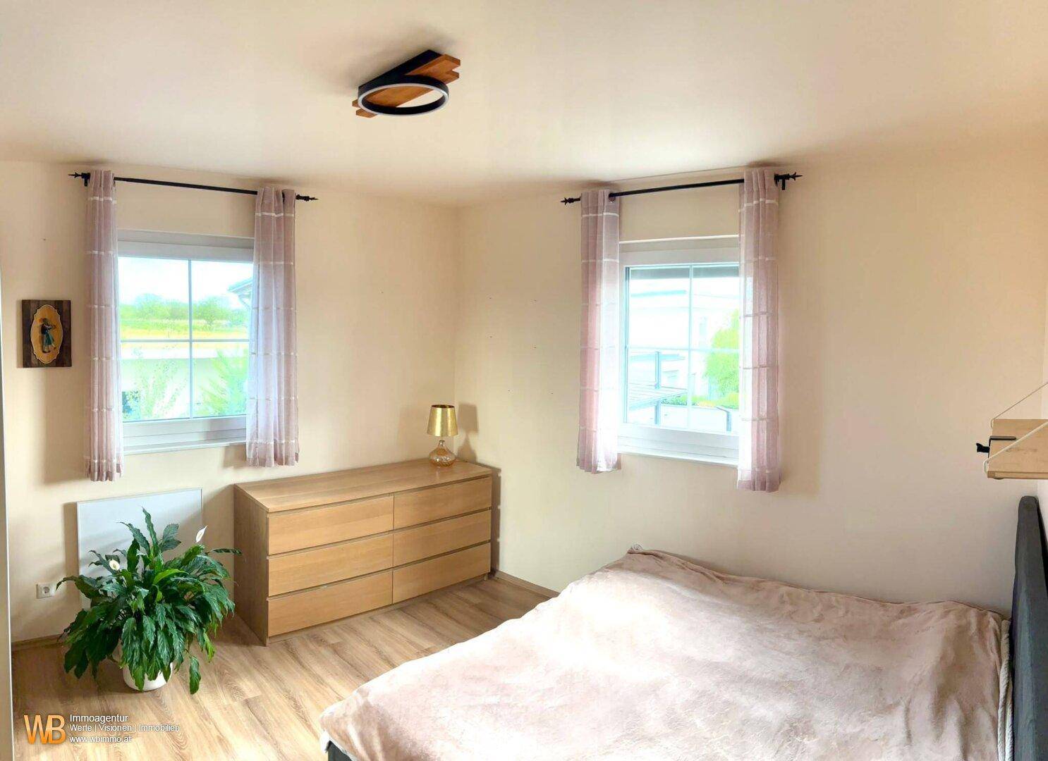 Helles Schlafzimmer mit zwei Fenstern, Holzboden und einfacher, funktionaler Einrichtung.