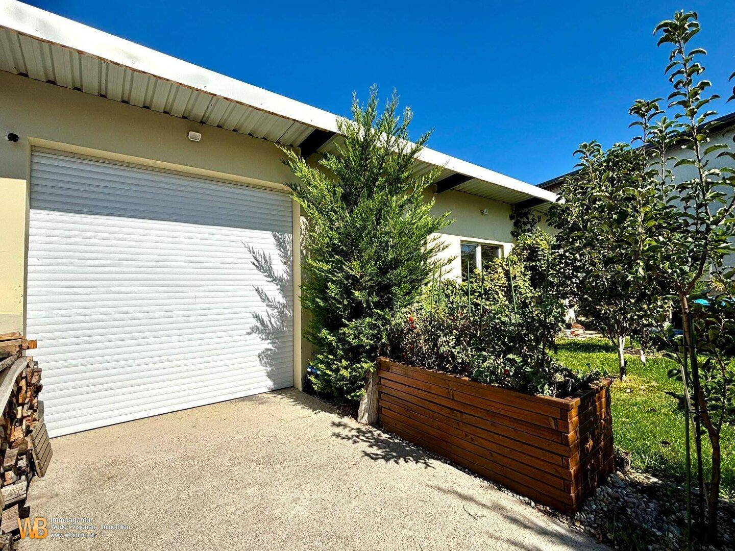Garage mit weißem Rolltor und angrenzendem, gepflegtem Gartenbereich mit Bäumen und Rasen.