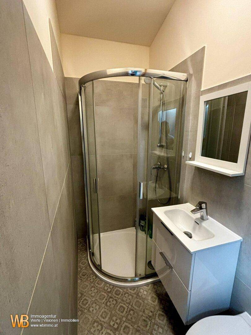 Kleines Badezimmer mit moderner Duschkabine, Waschbecken und grauen Wandfliesen.