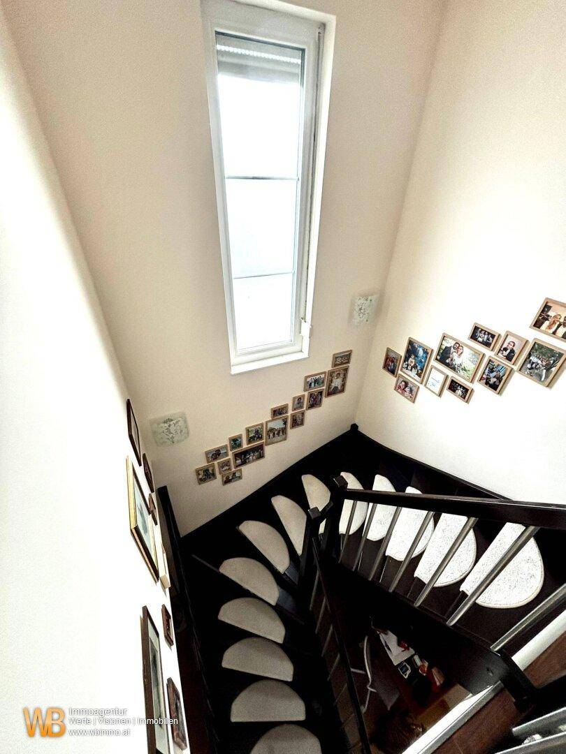 Helles Treppenhaus mit dunkler Holztreppe, hohen Wänden und einem hohen Fenster für Tageslicht.