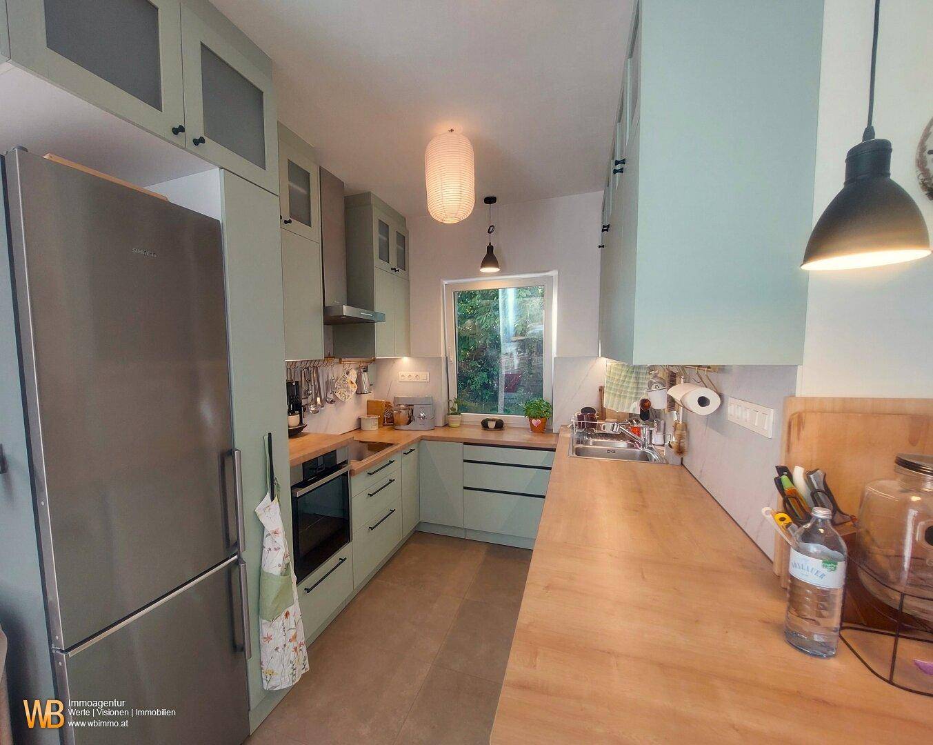 Moderne Küche in Mintgrün mit Holzarbeitsplatten, Edelstahlgeräten und Fenster zum Garten.