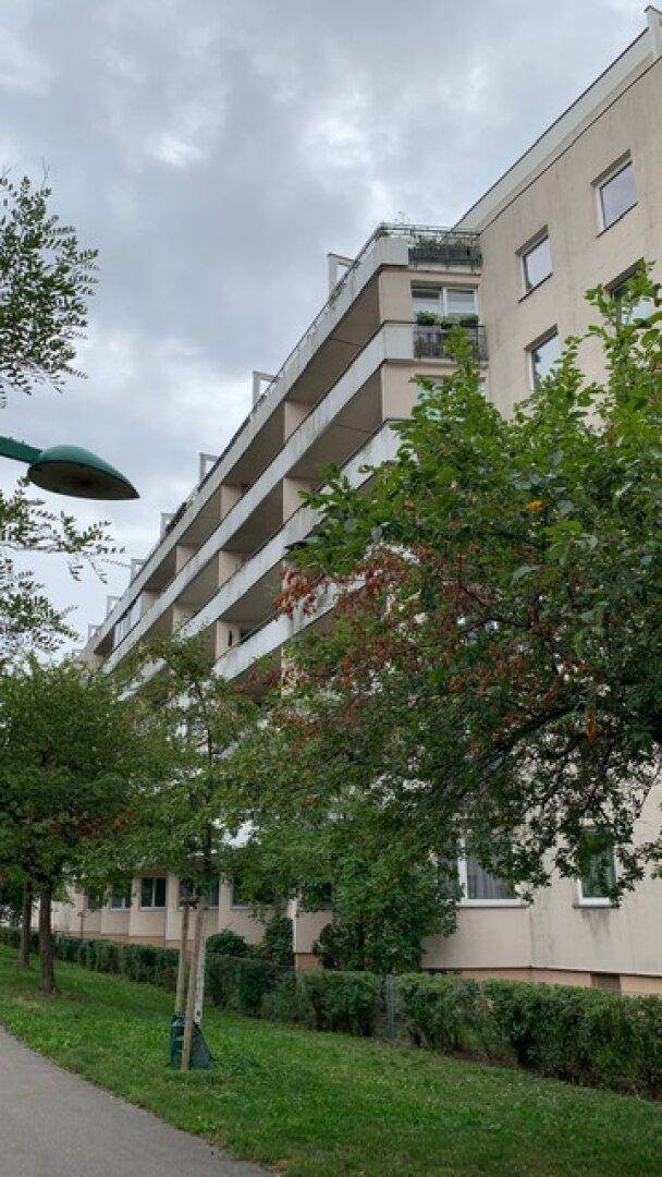 Ansicht eines Wohngebäudes mit mehreren Balkonen und umgebender Grünfläche unter bewölktem Himmel.