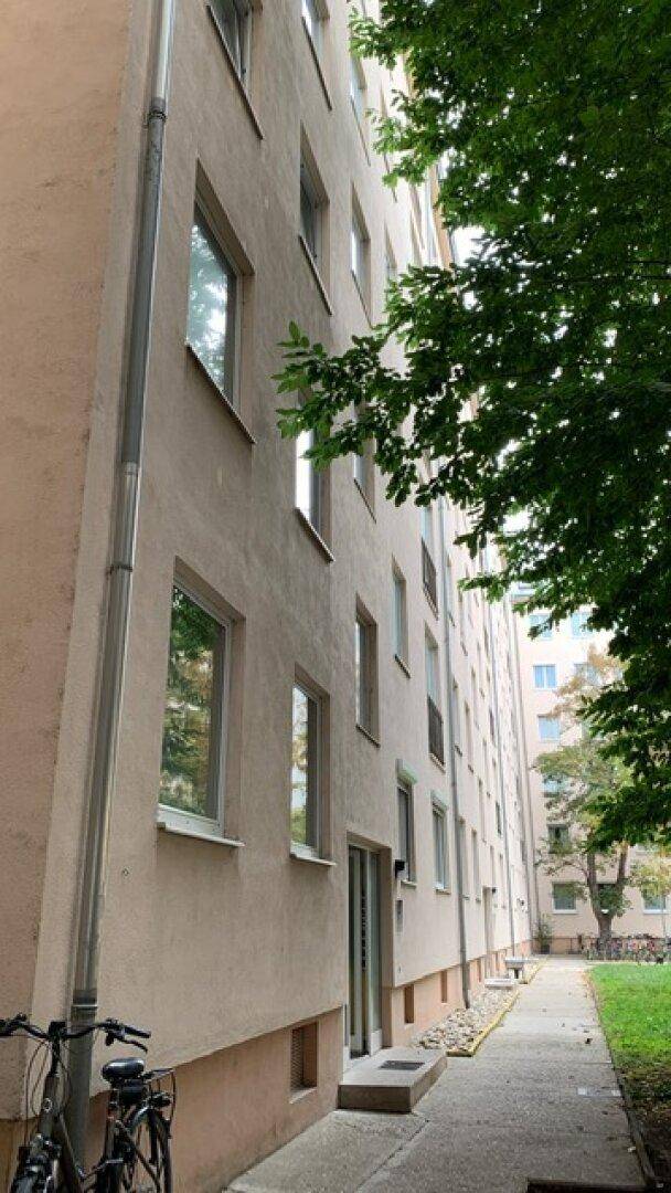 Die beige Fassade eines Mehrfamilienhauses mit zahlreichen Fenstern wird von Bäumen und einem Gehweg gesäumt.