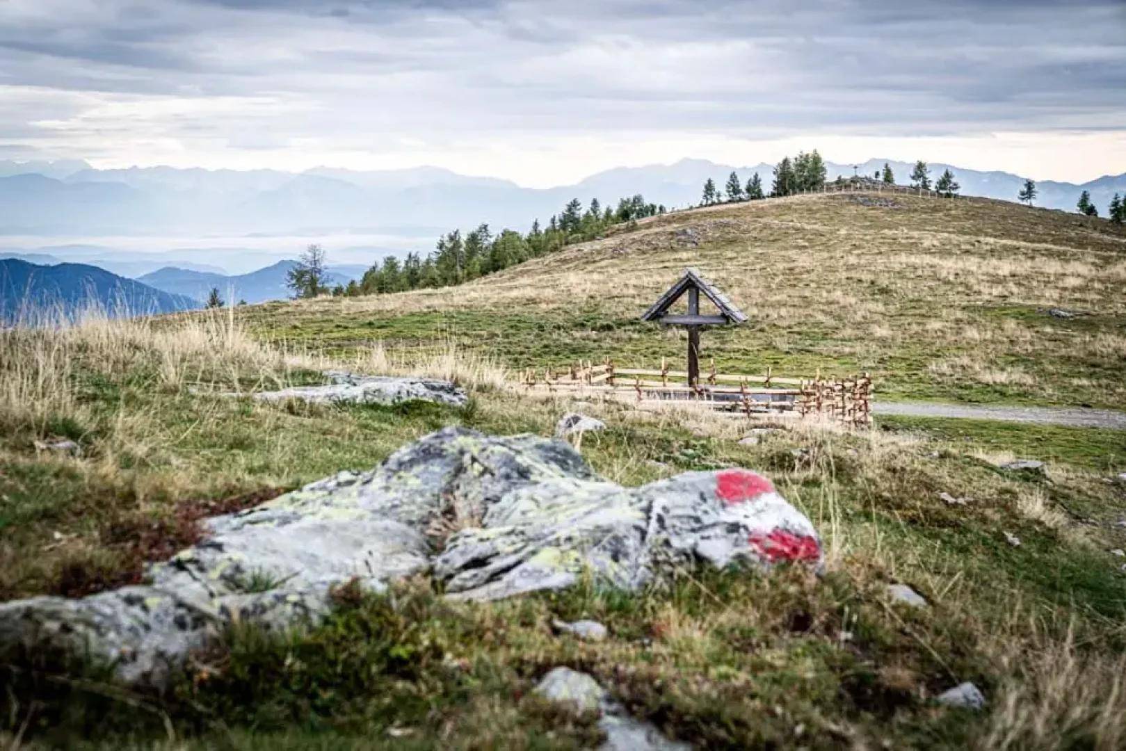 Hügelige Landschaft mit grasbewachsenen Hängen, einem Holzkreuz und Bergen im Hintergrund unter bewölktem Himmel.