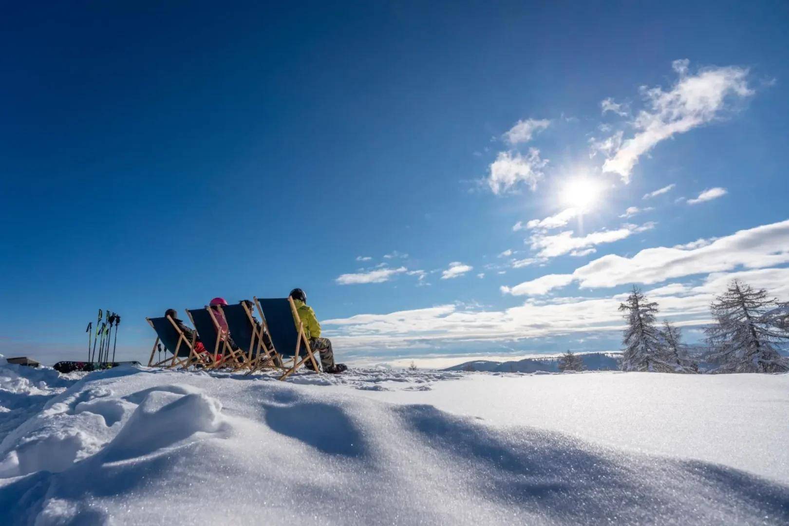 Menschen entspannen in Liegestühlen in einer weiten, schneebedeckten Landschaft unter strahlend blauem Himmel.
