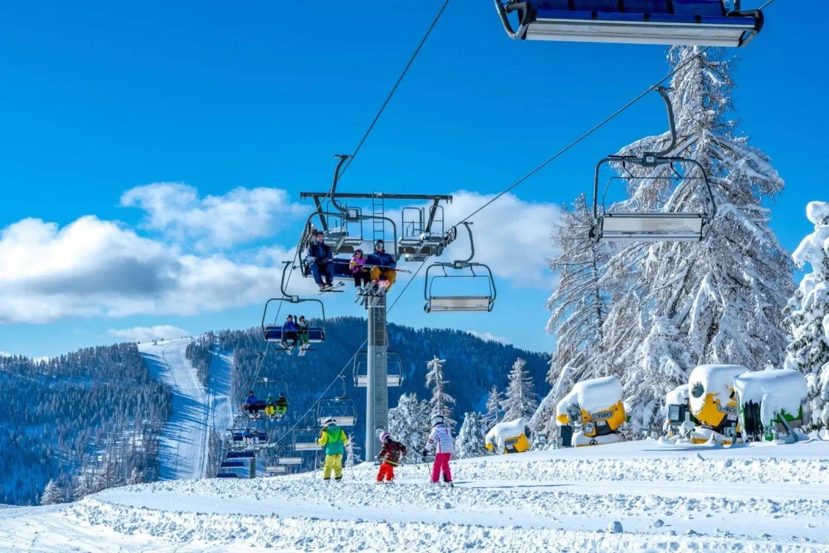 Skigebiet mit Skilift, der Skifahrer über schneebedeckte Pisten und verschneite Bäume unter blauem Himmel transportiert.
