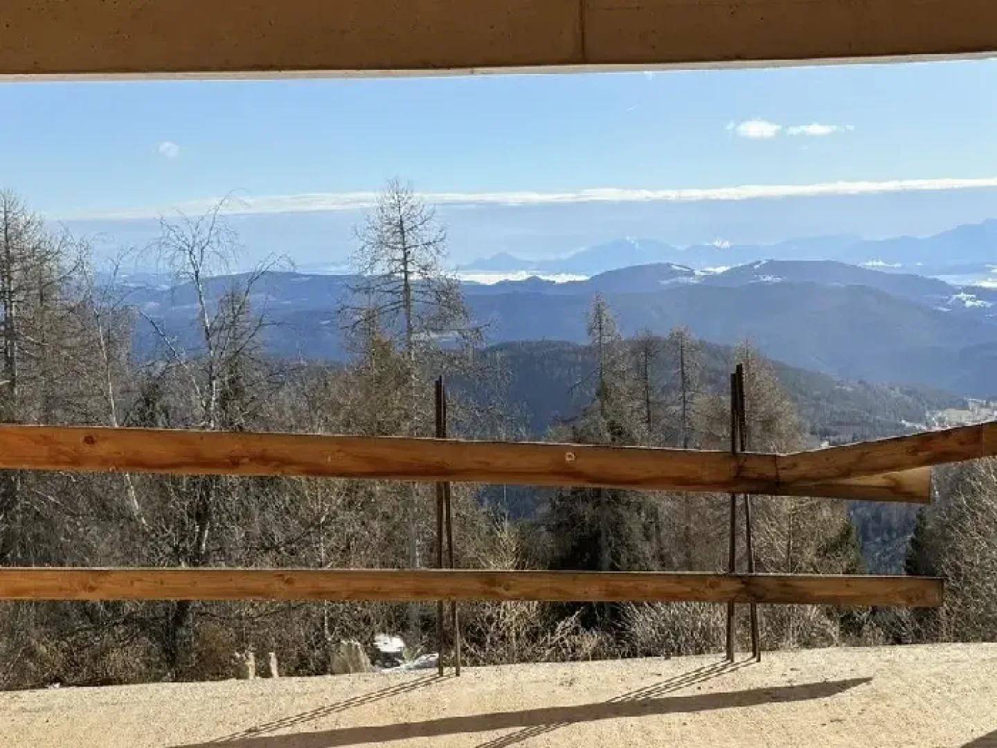 Rustikaler Holzzaun im Vordergrund mit einem weiten Blick auf die umliegende Berg- und Waldlandschaft.