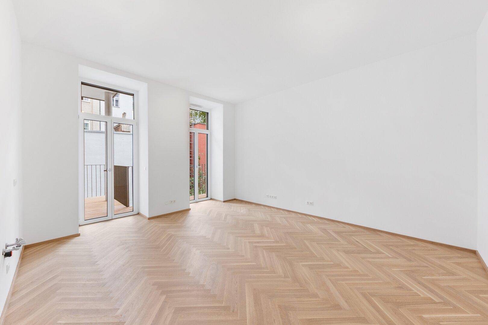 Lichtdurchfluteter Raum mit Fischgrätparkett und Zugang zu einem Balkon.