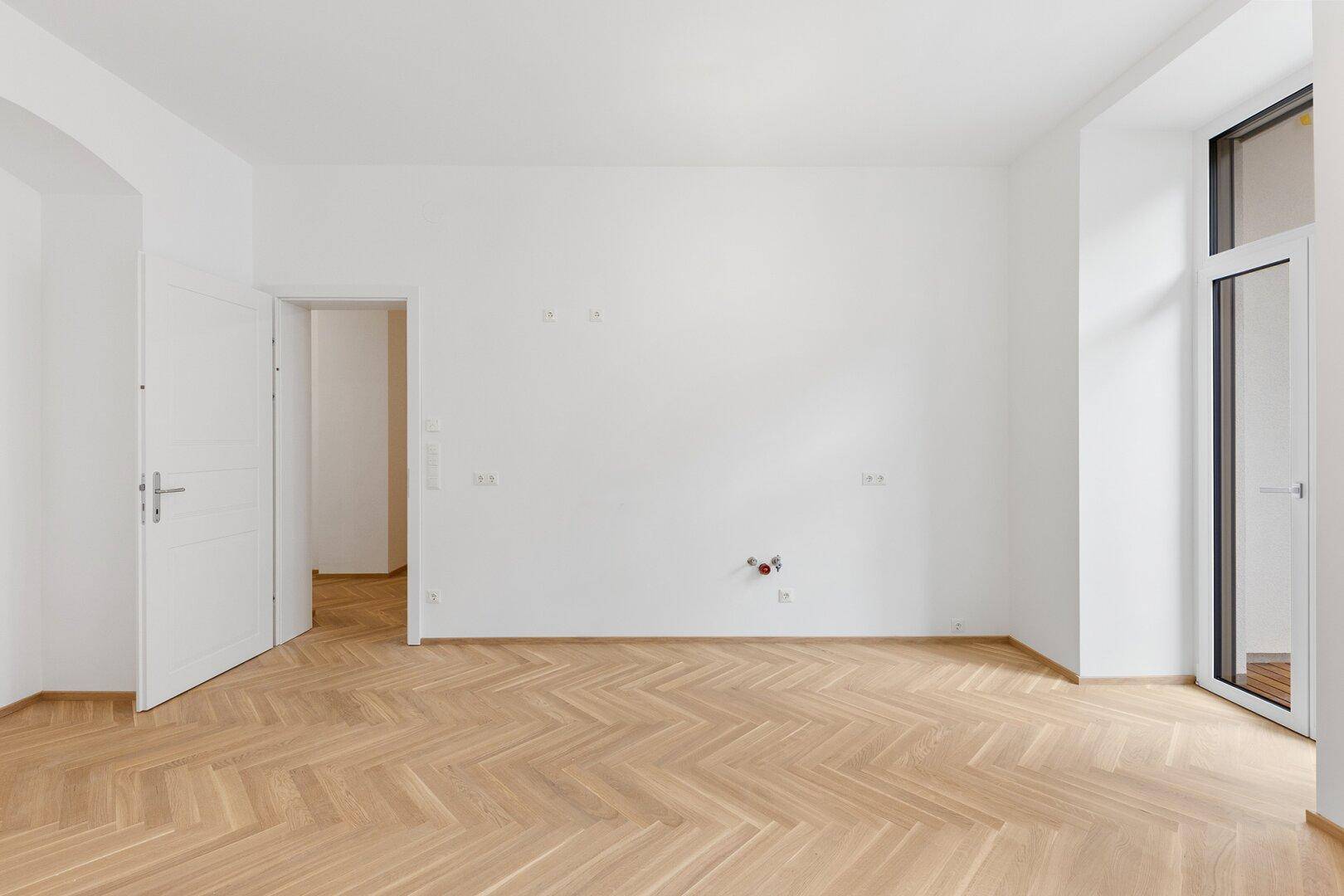 Heller Raum mit Fischgrätparkett und Zugang zu einem französischen Balkon.