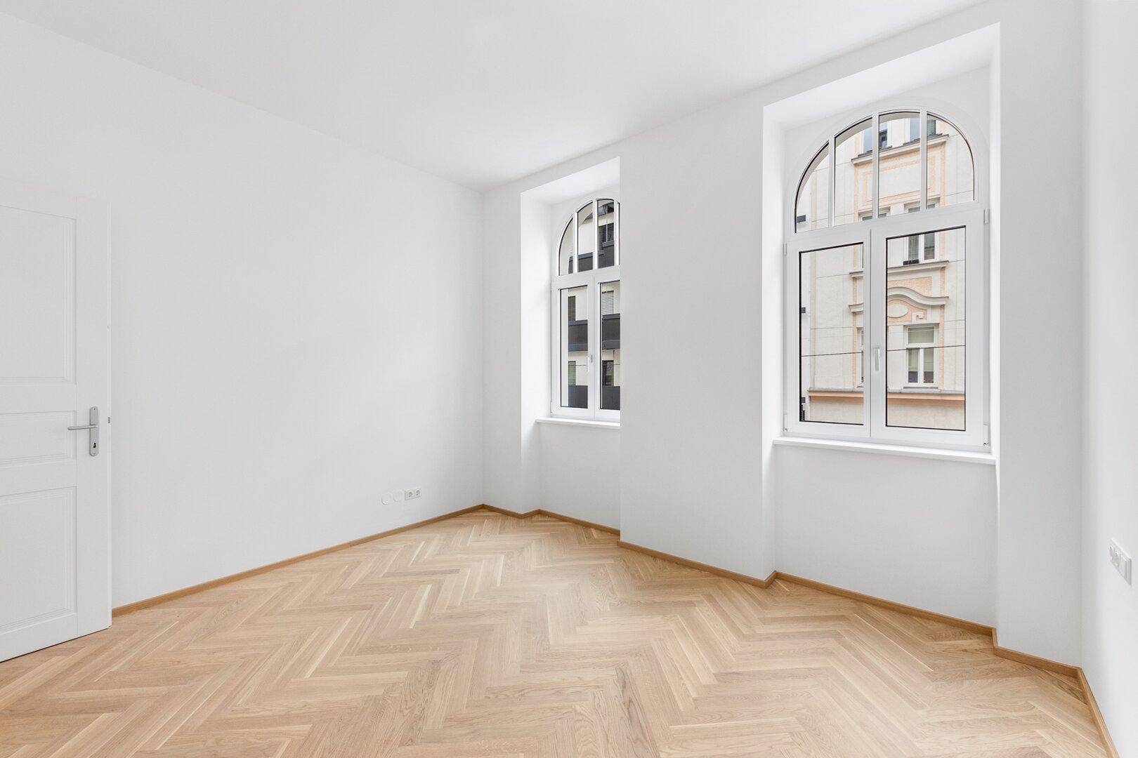Geräumiges Zimmer mit hellem Fischgrätparkett und zwei großen Rundbogenfenstern.
