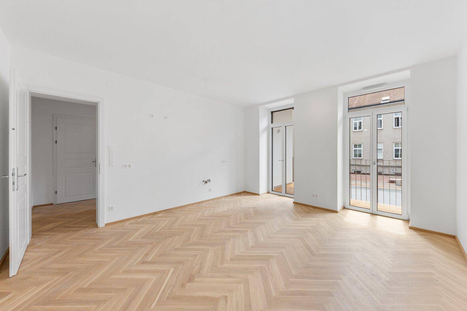 Lichtdurchflutetes Zimmer mit Fischgrätparkett und zwei Fenstertüren zum Balkon.