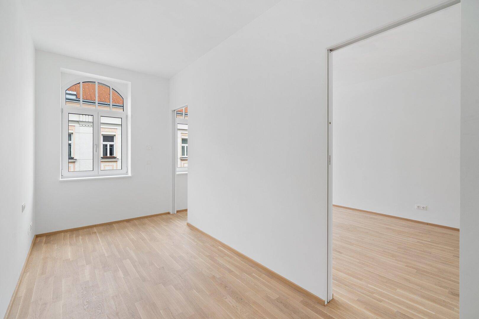 Heller, leerer Raum mit Holzfußboden, weißen Wänden und einem Bogenfenster, offen zu einem Nebenraum.