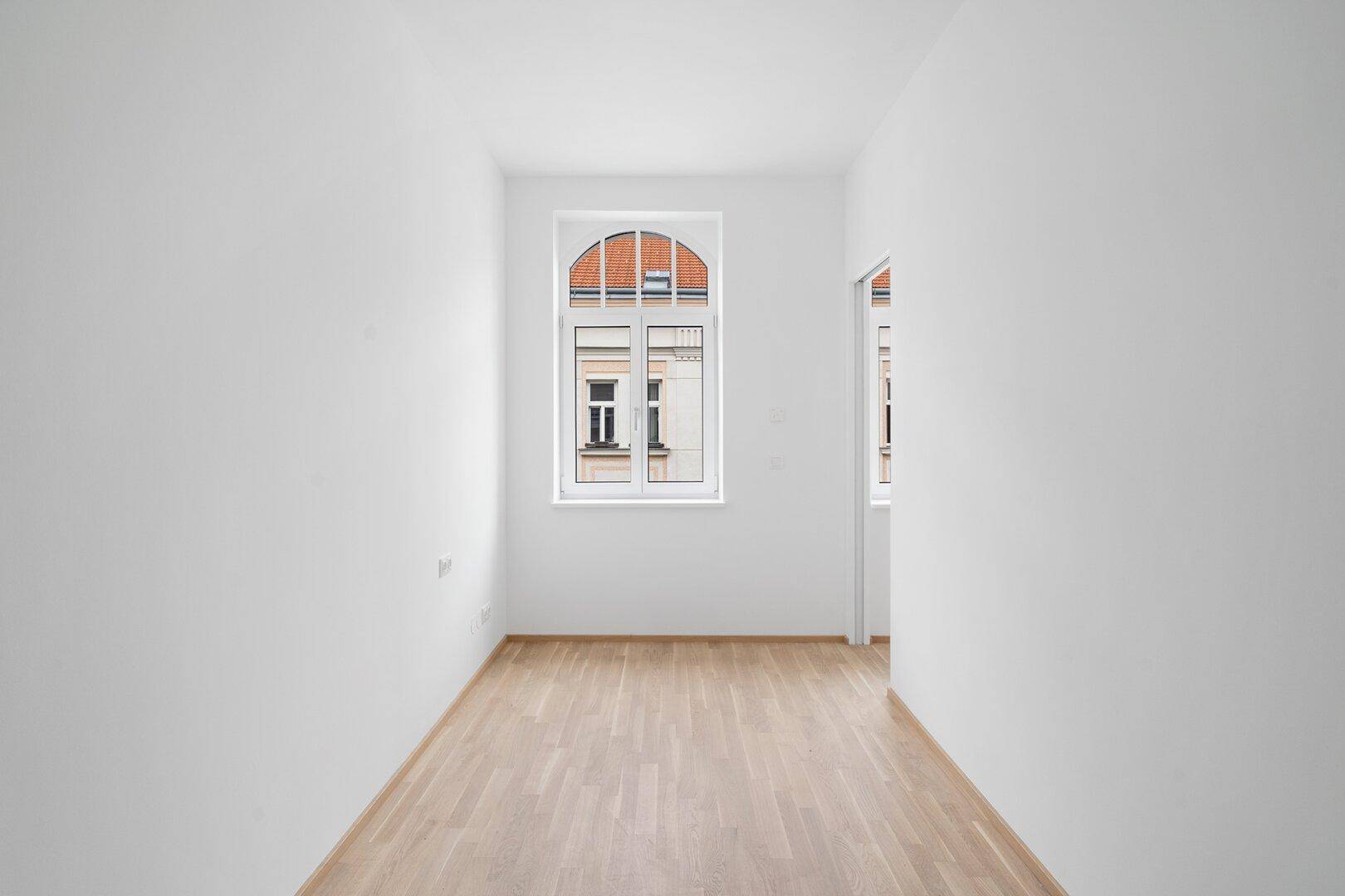 Leerer Raum mit hellem Holzfußboden, weißen Wänden und einem eleganten Bogenfenster.