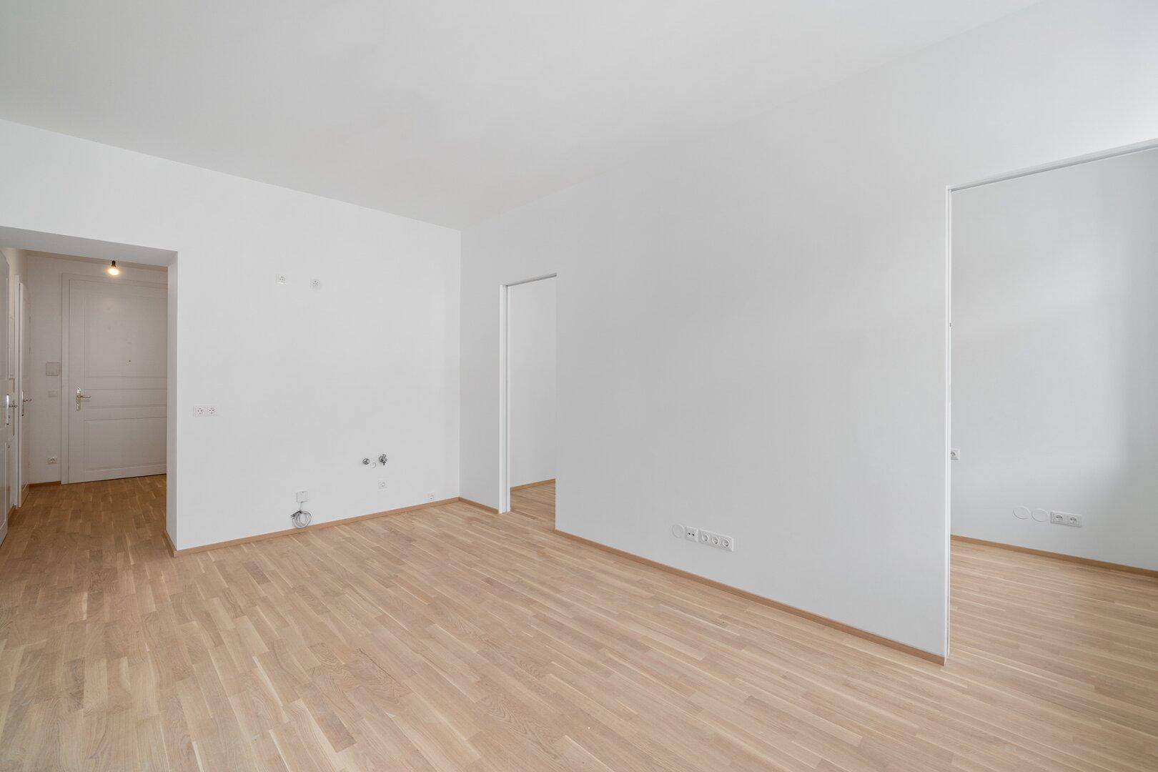 Geräumiger, leerer Raum mit hellem Holzfußboden und weißen Wänden, der mehrere Zugänge bietet.