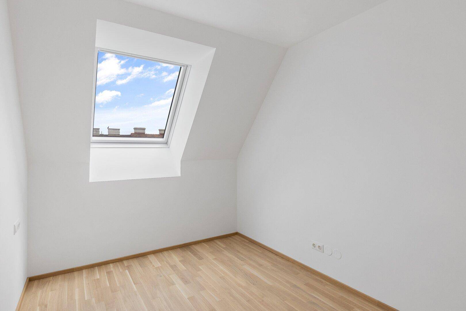 Gemütliches Dachzimmer mit einem großen Dachfenster, hellem Holzboden und weiß gestrichenen Wänden.