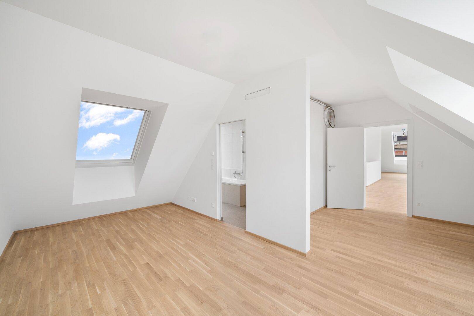 Heller Dachgeschossraum mit Parkettboden und Zugang zu einem angrenzenden Badezimmer.