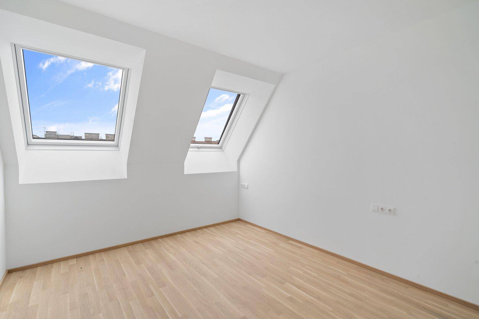 Heller Dachgeschossraum mit schrägen Wänden, zwei Dachfenstern und neuem Parkettboden.