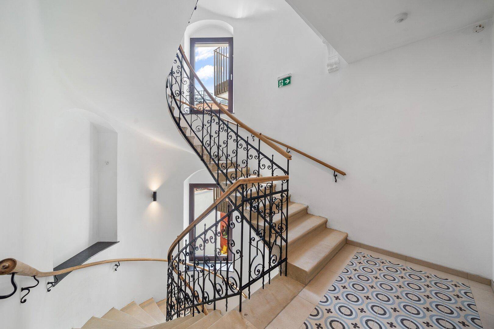 Geschwungene Treppe mit schmiedeeisernem Geländer und gemustertem Fliesenboden in einem historischen Treppenhaus.