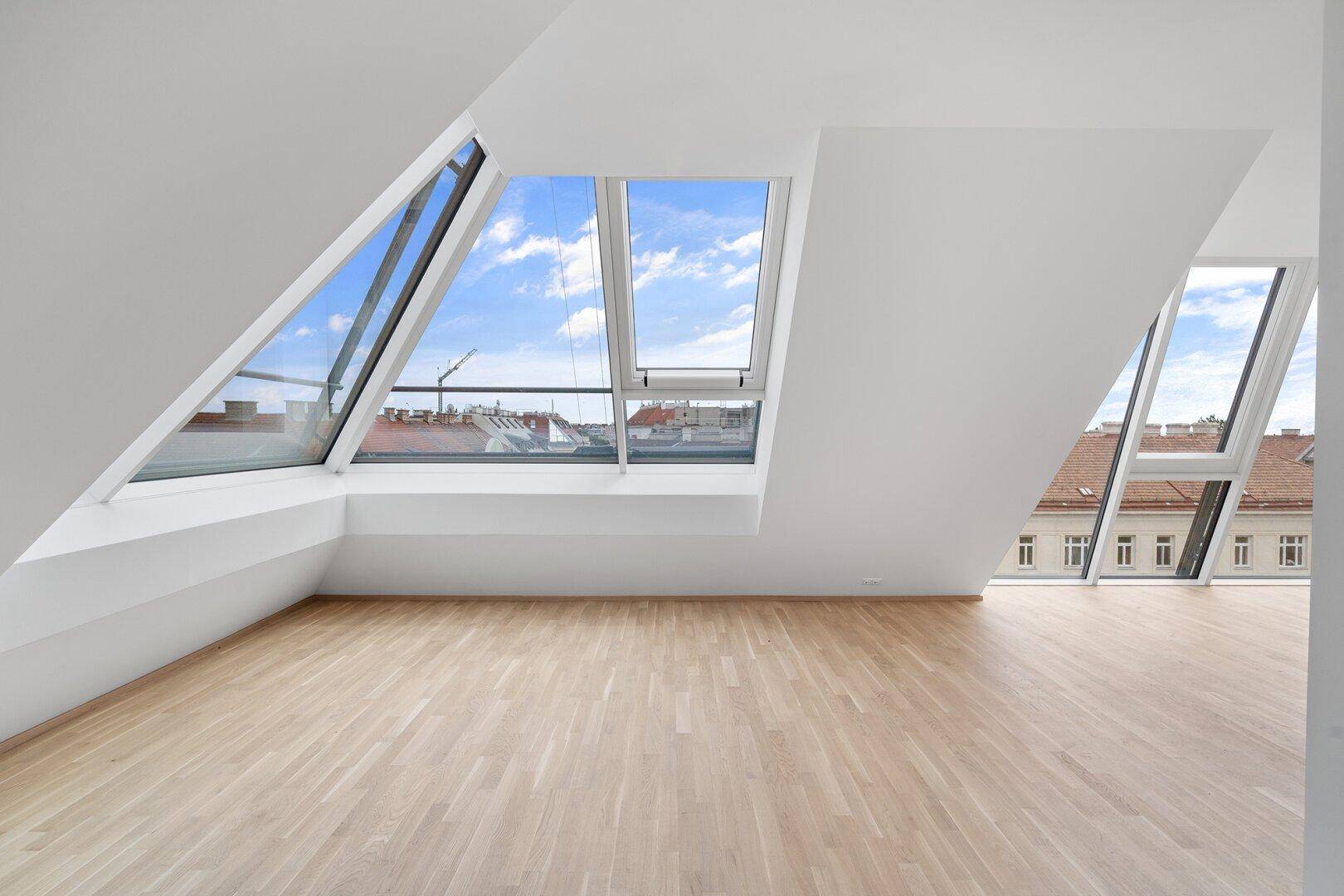 Heller Dachgeschossraum mit schrägen Wänden, großen Fenstern und Stadtblick.