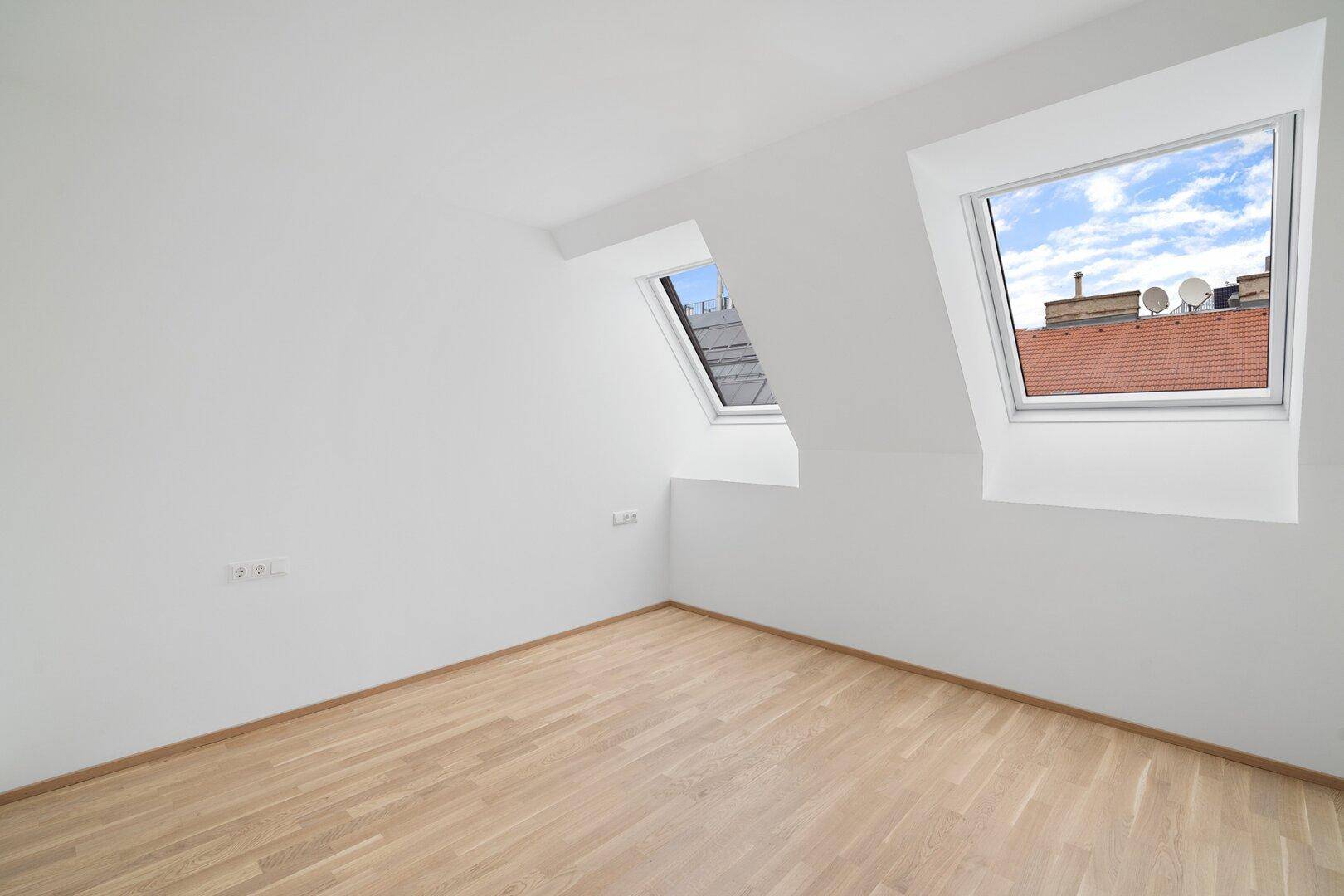 Dachzimmer mit Holzboden und zwei Dachfenstern, die viel Tageslicht hereinlassen.