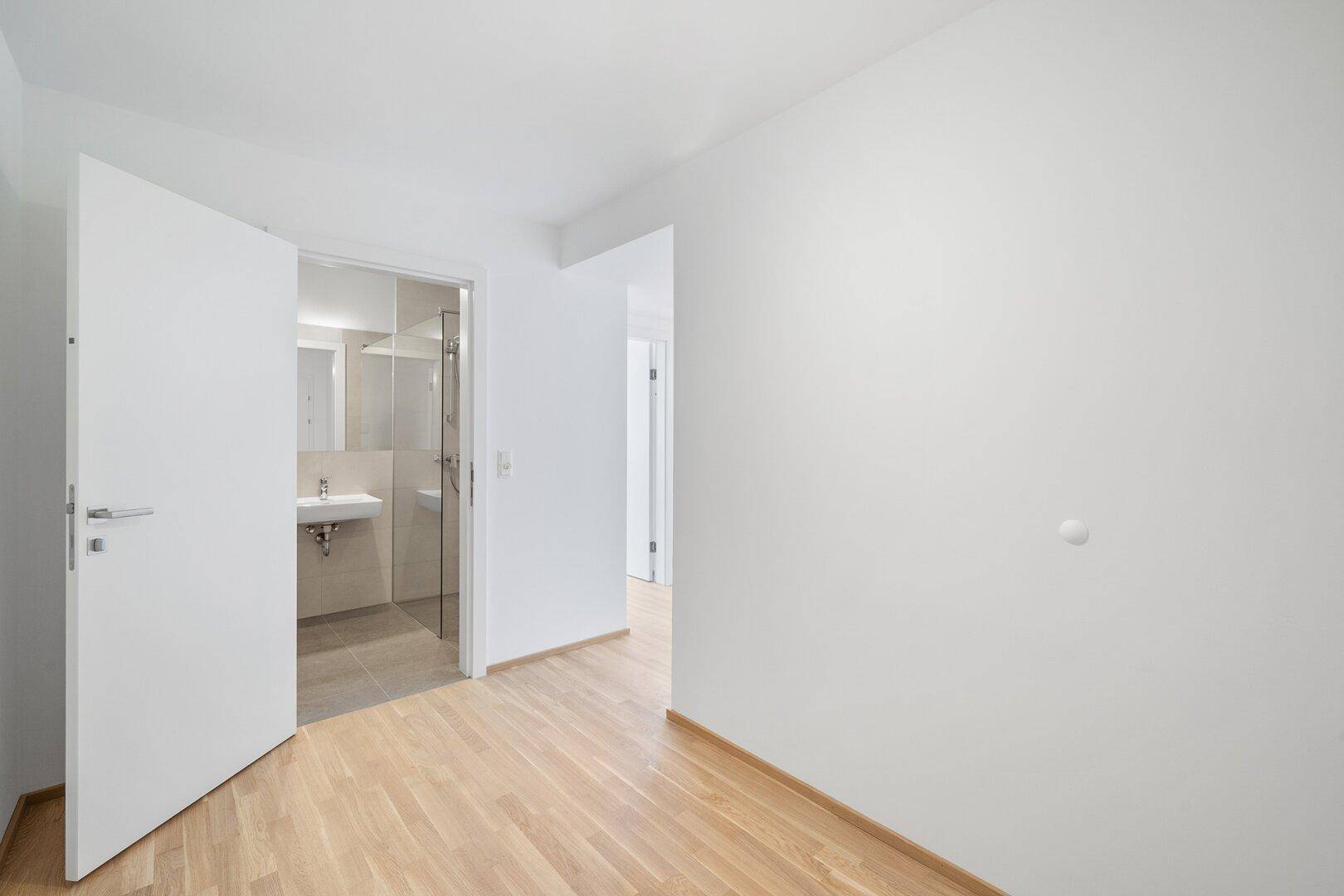 Helles Zimmer mit Holzboden und Zugang zu einem modernen Badezimmer mit Dusche.