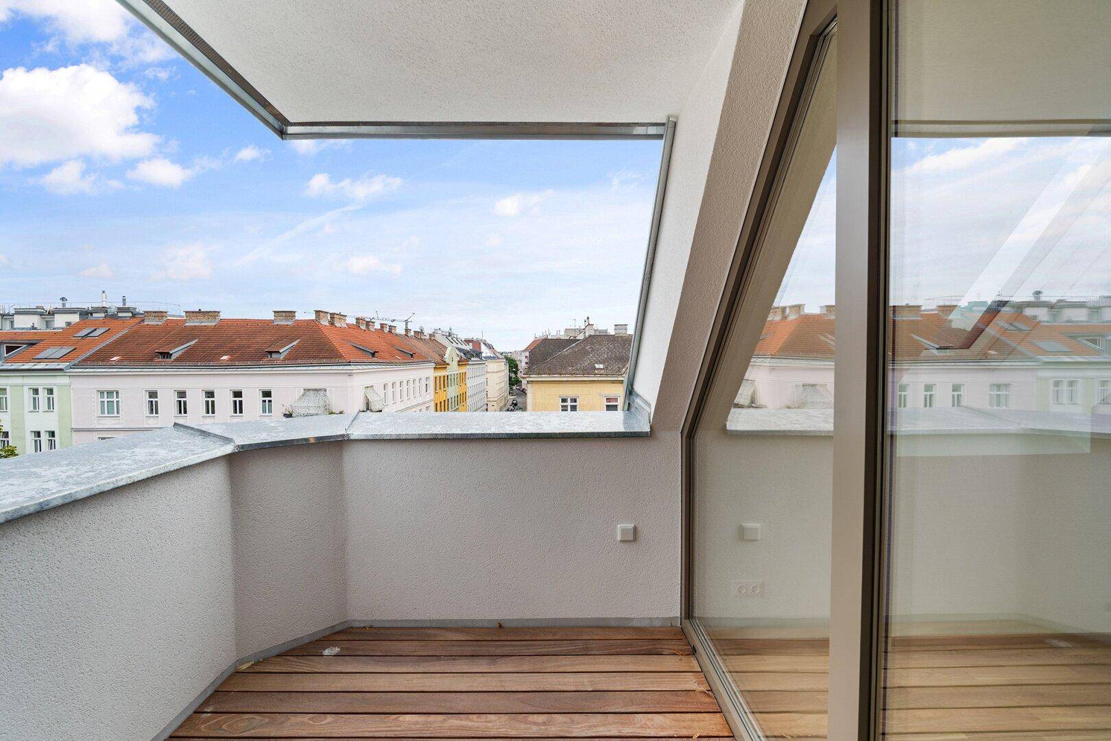 Kleine Loggia mit Holzdielen und Blick über die Dächer der Stadt unter blauem Himmel.