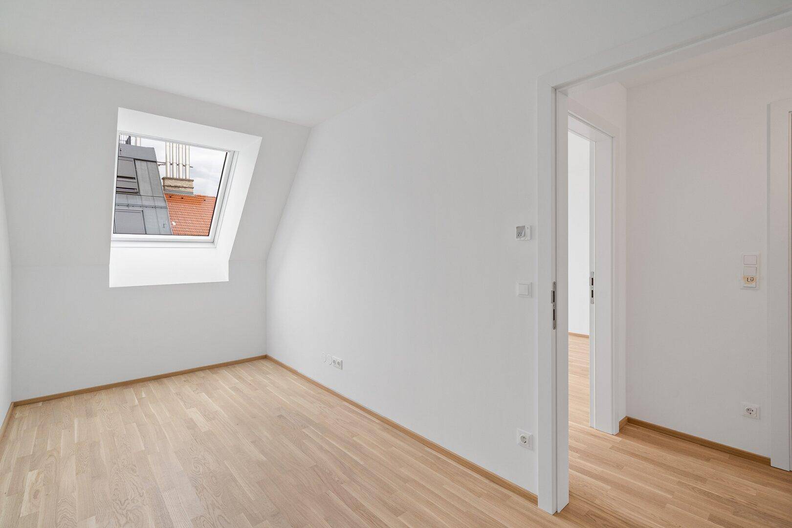 Helles Zimmer im Dachgeschoss mit Holzboden, weiß gestrichenen Wänden und Dachfenster.