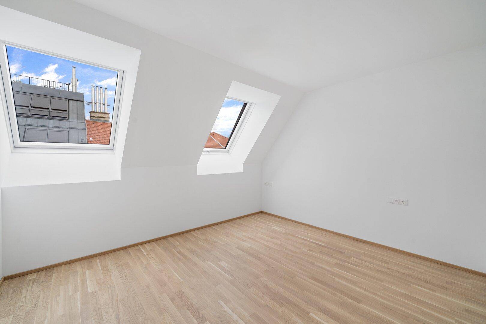 Lichtdurchflutetes Dachgeschosszimmer mit Holzboden, weißen Wänden und zwei Dachfenstern.