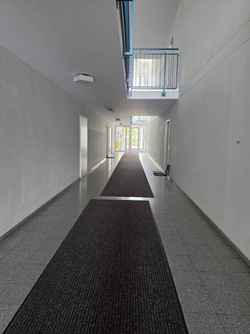 Langer, heller Flur mit grauem Fliesenboden und Blick auf einen Balkon am Ende.