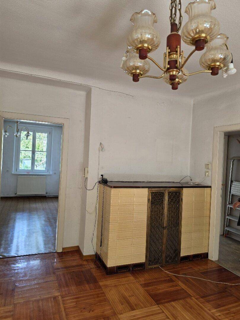 Geräumiges Zimmer mit traditionellem Kachelofen, Parkettboden und Zugang zu weiteren Räumen.
