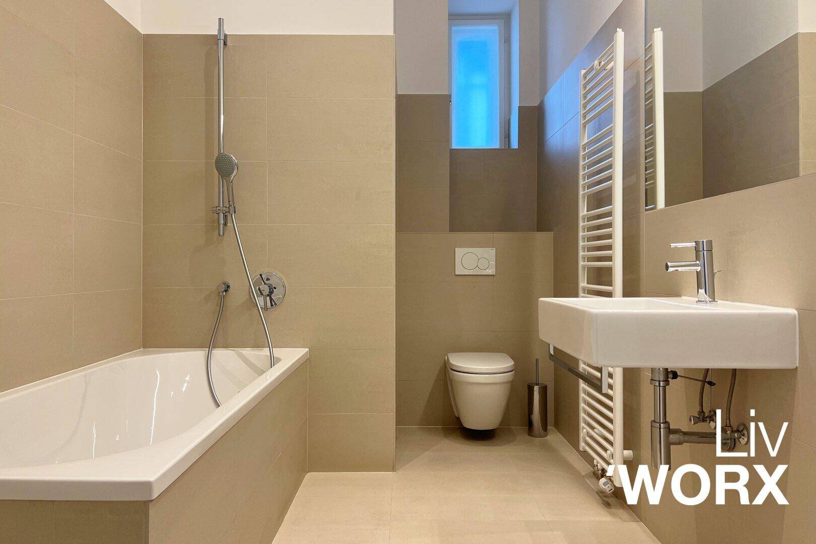 Modernes Badezimmer mit beigefarbenen Fliesen, Badewanne, wandhängendem WC und kleinem Fenster.