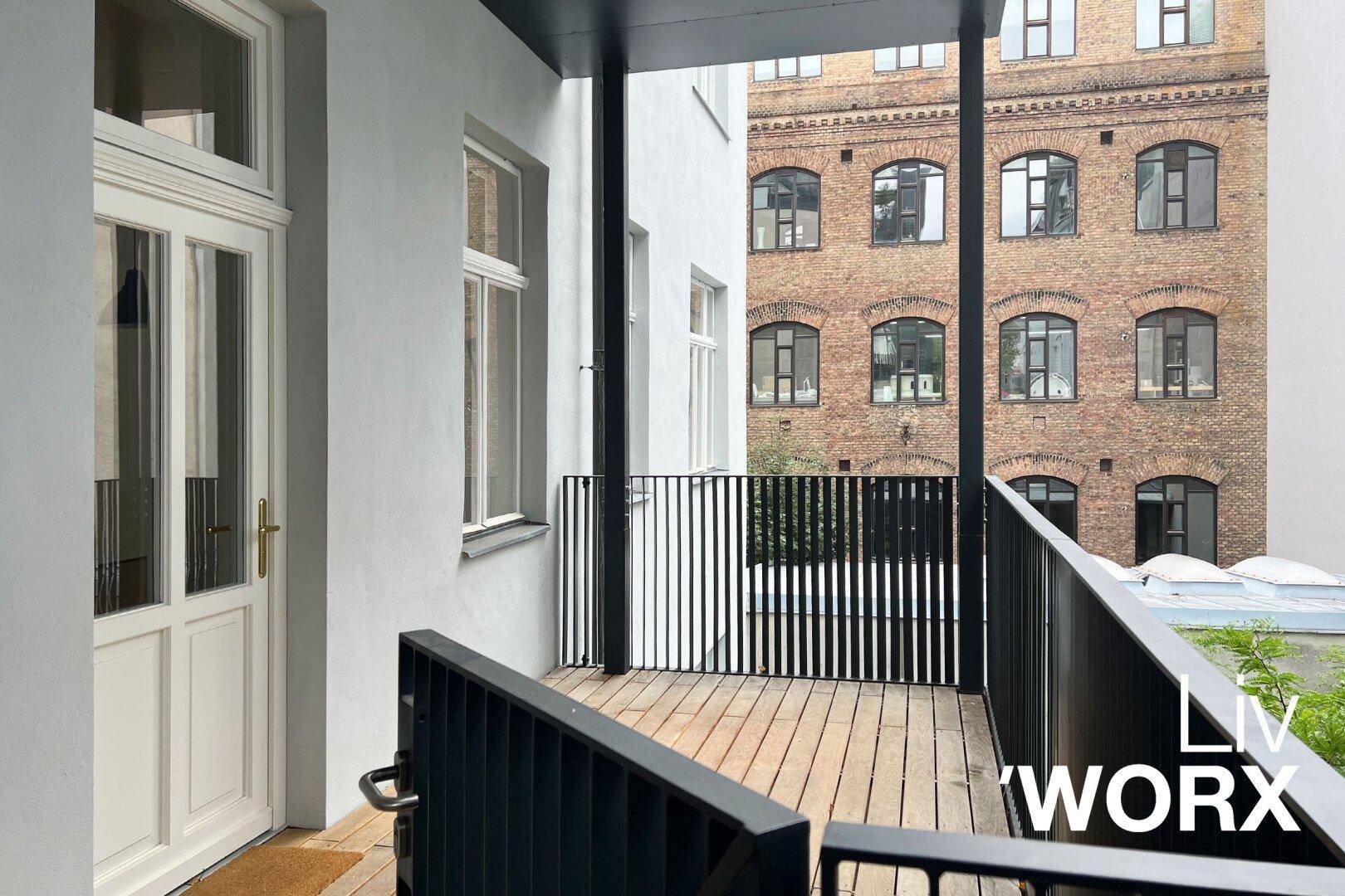 Kleiner Balkon mit Holzbelag und schwarzem Geländer, mit Blick auf ein Backsteingebäude.