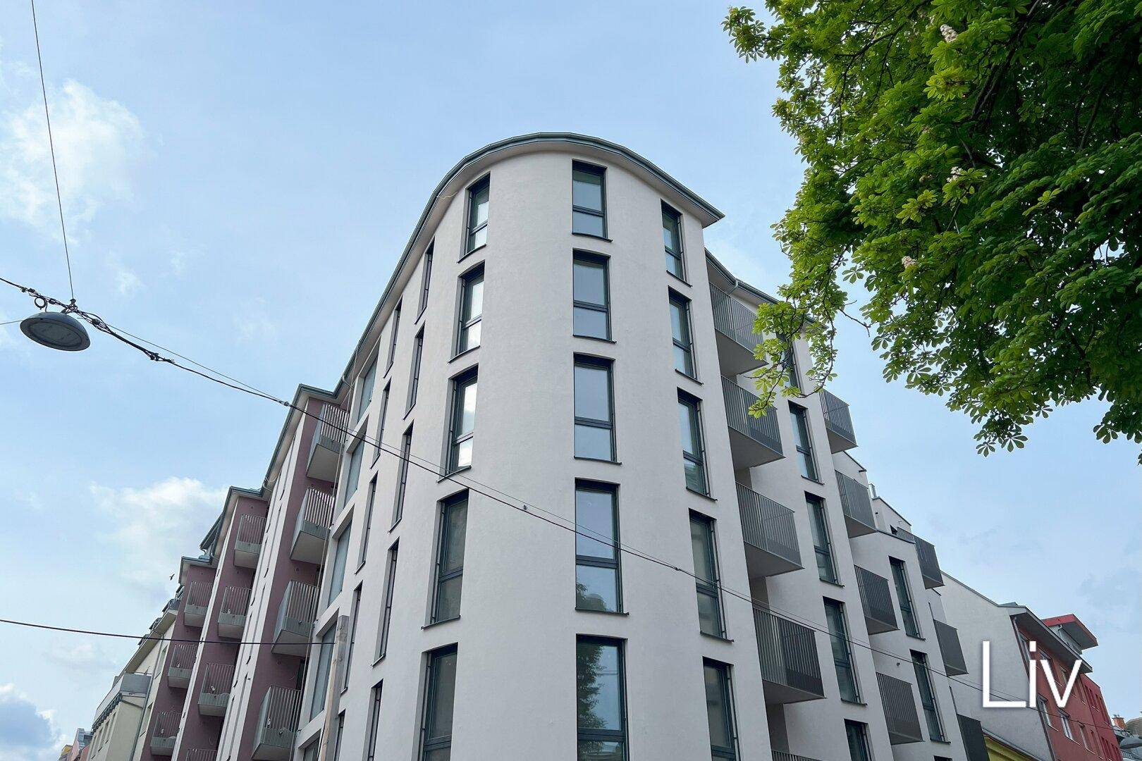 Gebäude mit moderner, geschwungener Fassade, vielen Fenstern und Balkonen, umgeben von Stadtgrün.