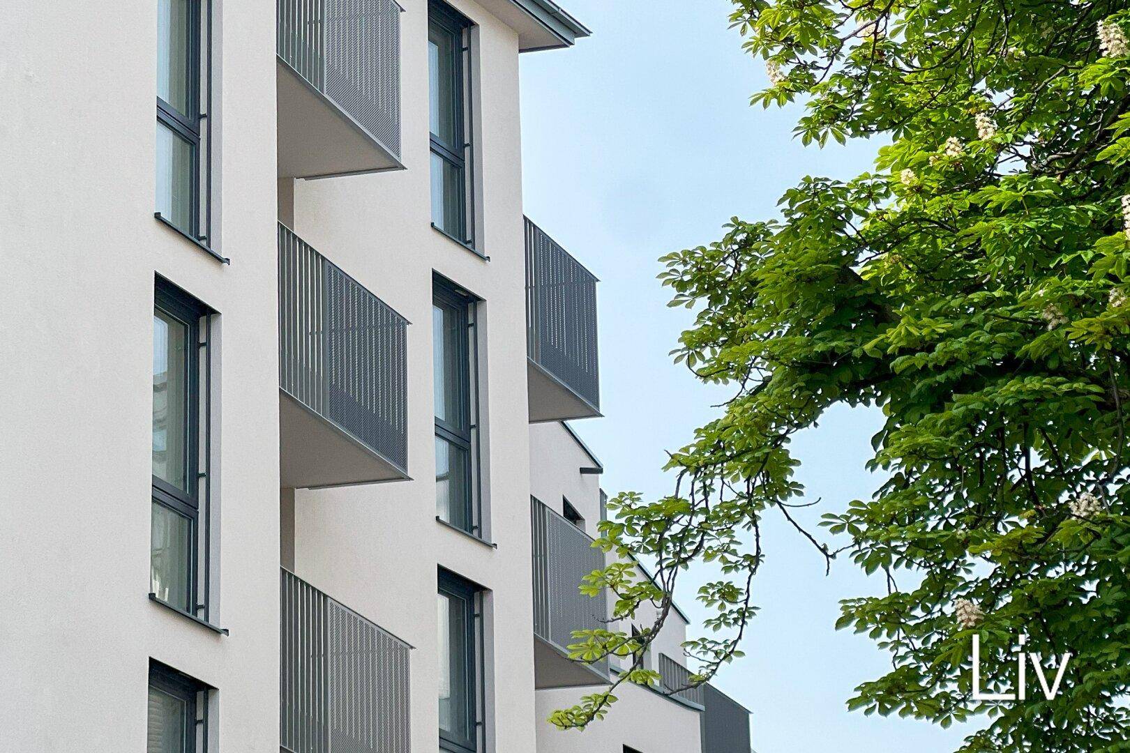 Moderne Fassade eines Wohngebäudes mit zahlreichen Balkonen und großen Fenstern, umgeben von grünen Bäumen.