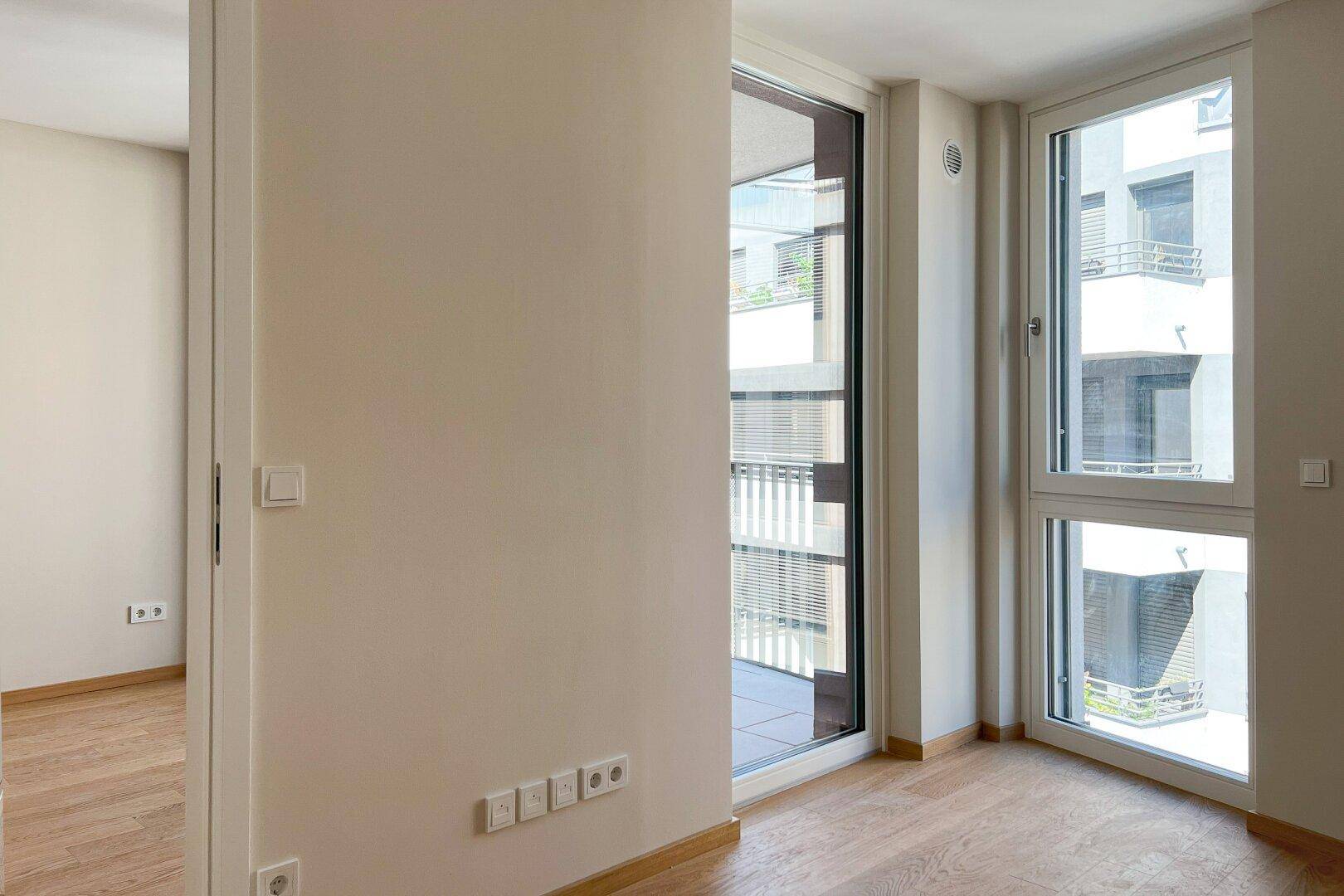 Heller, leerer Raum mit Holzboden und großem Fenster mit Zugang zu einem Balkon.