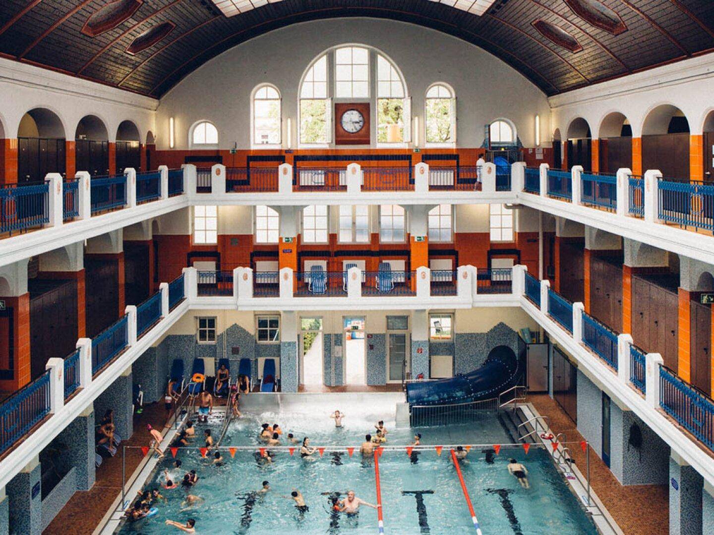 Großes historisches Hallenbad mit vielen Schwimmern, hohen Decken und viel natürlichem Licht.