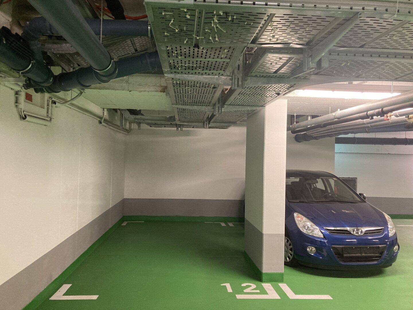 Ein Stellplatz in einer Tiefgarage mit einem blauen Auto, sichtbaren Rohren und Kabeltrassen.