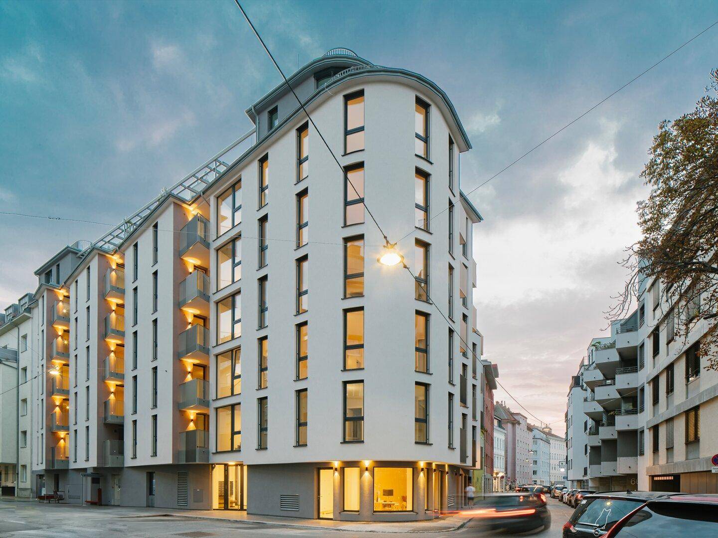 Modernes Eckgebäude mit vielen Fenstern, Balkonen und einer Dachterrasse in urbaner Umgebung.