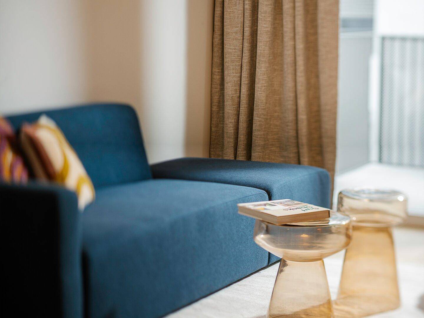 Detailansicht eines modernen blauen Sofas mit stilvollen Beistelltischen und hellen Vorhängen am Fenster.