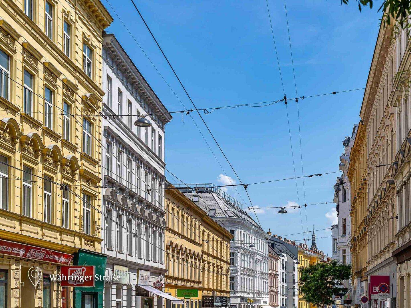Belebte Straße mit historischen Gebäuden und Geschäften unter blauem Himmel.